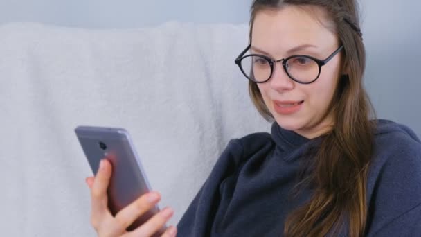 Glückliche junge brünette Frau mit Brille spricht einen Videochat auf einem Handy im Sessel sitzend. — Stockvideo