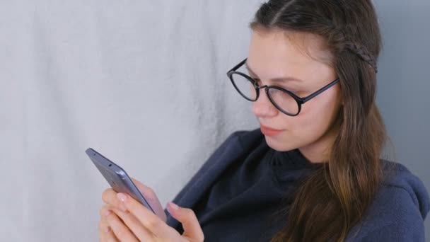 Joven morena en gafas escribe un mensaje en un teléfono móvil sentado en sillón en casa . — Vídeos de Stock