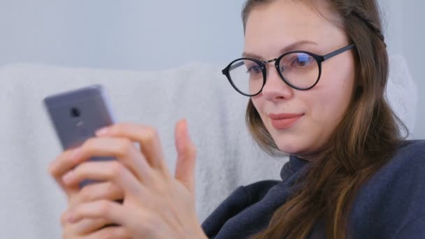 Feliz joven morena en gafas de hablar de un chat de vídeo en un teléfono móvil sentado en un sillón . — Vídeos de Stock