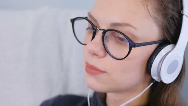 Молода жінка в окулярах і навушниках слухає музику, обличчя крупним планом . — стокове відео