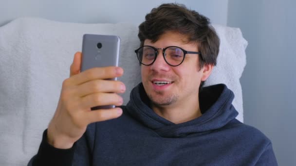 Šťastný mladý muž v brýlích mluvící video chatu na smartphone v křeslo, online data. — Stock video
