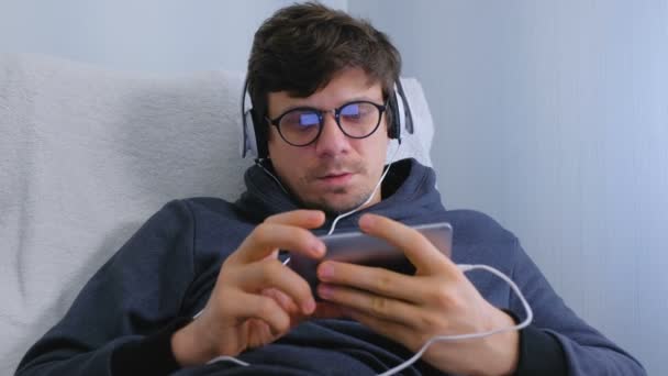 Hombre en gafas y auriculares de navegación en línea en el teléfono inteligente. Primer plano de la cara . — Vídeo de stock