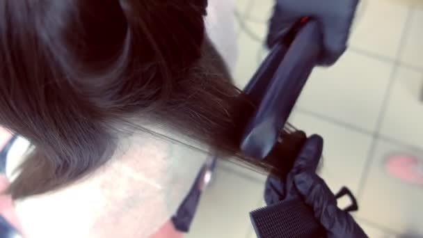 Hairdresser straights dark brown hair of beautiful woman using hair tongs in beauty salon. Hair close-up. — Stock Video