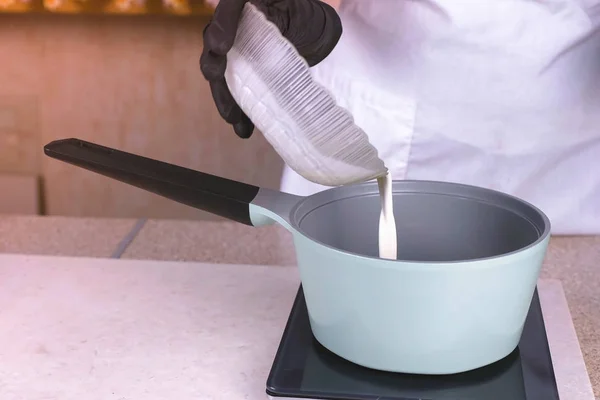 Cozinhe derrama o creme da tigela na panela para reaquecer. Mãos em luvas de borracha preta close-up . — Fotografia de Stock