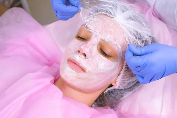 Cosmetologist puts a plastic film on top of the moisturizing mask on the womans face. — Stock Photo, Image