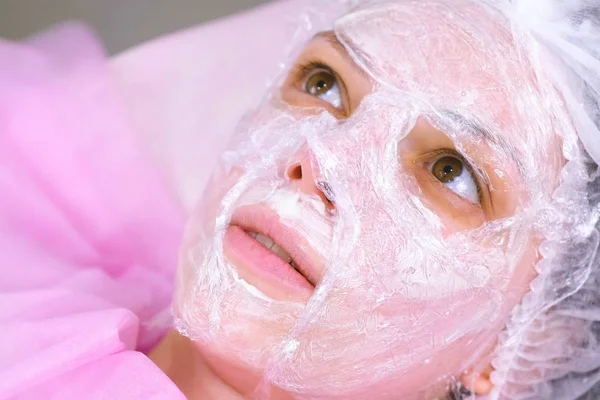 Cosmetólogo con una película de plástico en la parte superior de la máscara hidratante en la cara de las mujeres . — Foto de Stock