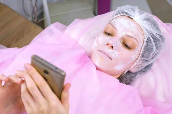 Женщина читает что-то в мобильном телефоне, лежащем на процедуре в офисе косметолога с маской на лице . — стоковое фото