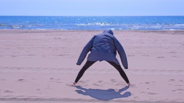 Frau in Jacke wärmt sich am Strand auf und streckt die Beine, Rückenansicht.