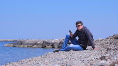 Man gezgin deniz taş plajda oturur, gülümsüyor ve akıllı telefon mesajları türleri.