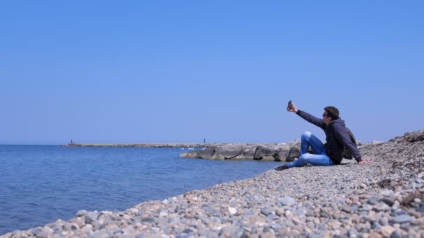 Man traveller is sitting on sea stone beach and making selfie on smartphone. — Stock Video