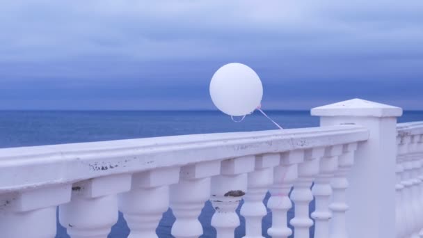 Bílý balon ve větru přivázaný k plotu na mořském nábřeží za oblačného dne. — Stock video