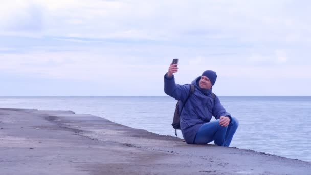 男人旅行者在智能手机上自拍坐在海边的旧海滨在冬天. — 图库视频影像