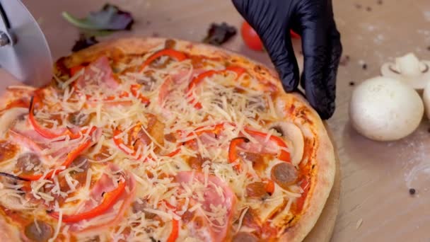 Кук режет пиццу с ветчиной, грибами и перцем на деревянной доске на столе. Руки крупным планом в резиновых перчатках . — стоковое видео
