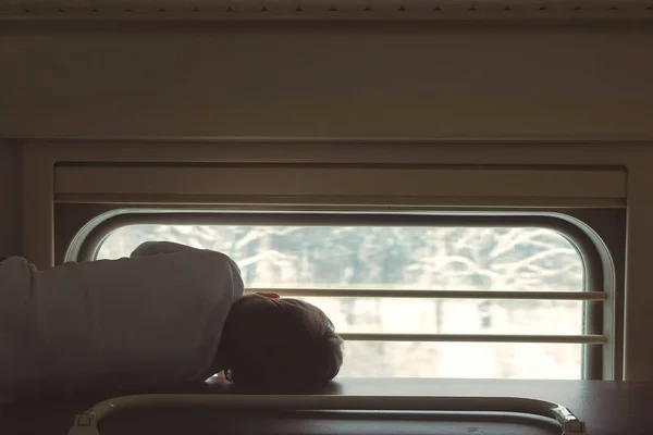 Rapaz no comboio deitado na prateleira de cima numa carruagem de segunda classe e a olhar para a janela. Visão traseira . — Fotografia de Stock