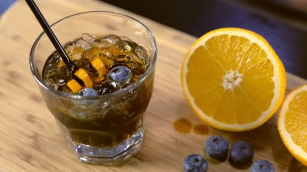 Cocktail alcolico sul bicchiere roccioso con arancia, ghiaccio e mirtillo sul tavolo, Vista laterale . — Video Stock