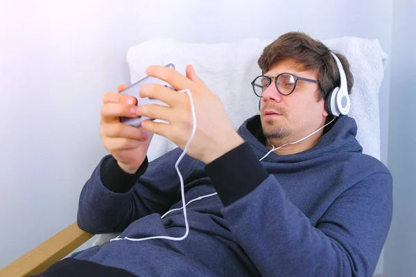 Чоловік в окулярах і навушниках грає в гру в смартфоні, сидячи в кріслі. Гамер розслабляється . — стокове фото