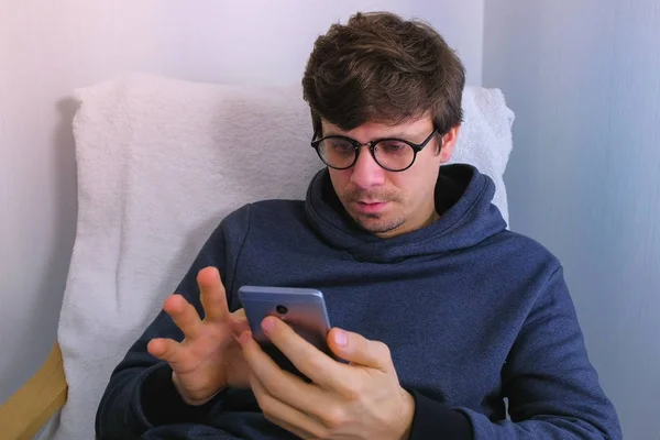 Człowiek w okularach jest przeglądanie w social media na smartfonie, siedząc w fotelu. — Zdjęcie stockowe