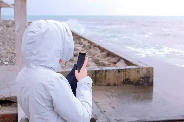 Mujer con una chaqueta blanca toma un teléfono móvil ondas de tormenta de pie en el terraplén. Vista trasera . —  Fotos de Stock