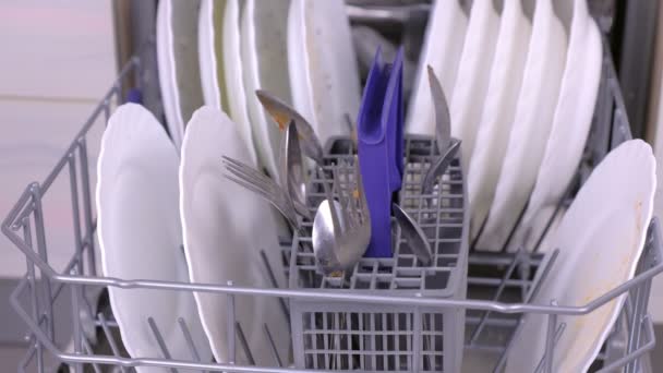 Манная рука кладет в посудомоечную машину белые грязные тарелки и ложки и запускает ее на кухне . — стоковое видео