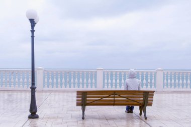 Kadın beyaz ceket ve hood sahildeki deniz manzaralı güzel terasta bankta oturuyor. Arkadan Görünüm.