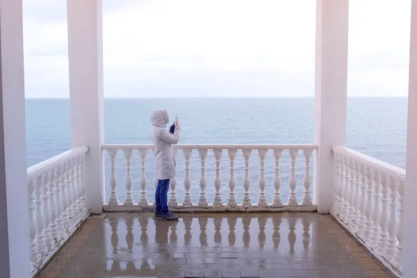 Mujer en la chaqueta blanca dispara vídeo olas del mar en el teléfono inteligente en la hermosa terraza con vista al mar. Vista trasera . —  Fotos de Stock