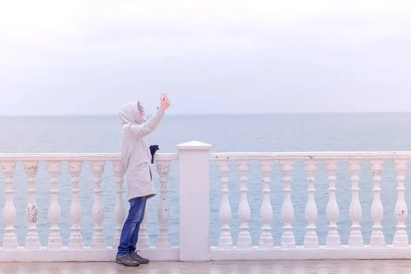 Femme en veste blanche et capuche fait selfie vidéo sur smartphone sur belle terrasse avec vue sur la mer sur le front de mer. Vue latérale . — Photo