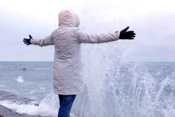 Kobieta w białej kurtce stoi na nabrzeżu na plaży patrzy na fale burzy. Widok z boku. — Zdjęcie stockowe