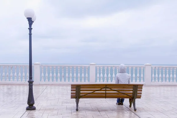 Femme en veste blanche et capuche est assise sur le banc sur une belle terrasse avec vue sur la mer sur le front de mer. Vue arrière . — Photo