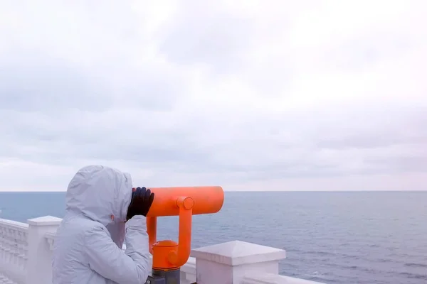 Femeia cu jachetă albă și capotă se uită în binocluri la malul mării cu vedere la mare. Vedere laterală . — Fotografie, imagine de stoc