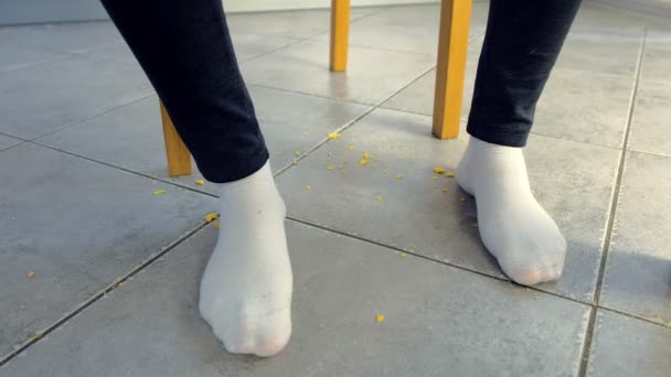 女性はテーブルで食べ、床にコーンフレークを砕きます。脚と床のクローズアップ、正面図. — ストック動画