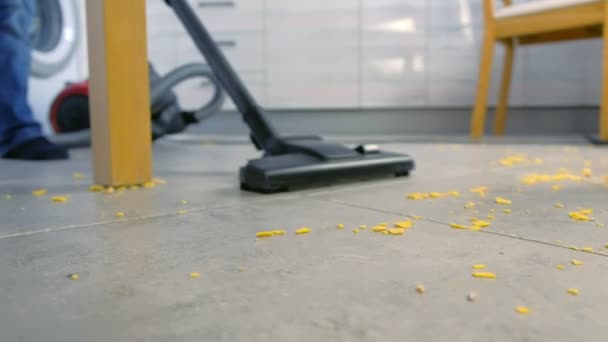台所の床を掃除する子供。彼は灰色のタイルに散らばったトウモロコシのフレークを片付けます。脚と掃除機クローズアップ. — ストック動画