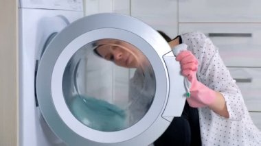 Pembe kauçuk eldiven kadın zemin üzerinde oturan, bez ile çamaşır makinesi yıkıyor. Yan görünüm, yakın çekim.
