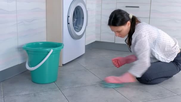 Mujer cansada en guantes de goma rosa lava y frota duro la mancha en el suelo de la cocina con un paño. Azulejos grises en el suelo . — Vídeo de stock