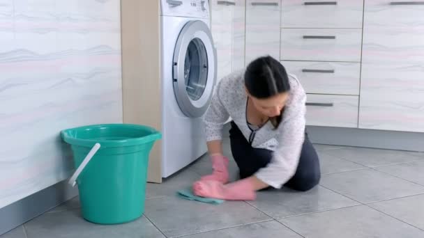 Mujer cansada en guantes de goma rosa lava el suelo de la cocina con un paño y mira a la cámara. Azulejos grises en el suelo . — Vídeo de stock