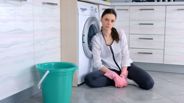 Mulher cansada em luvas de borracha rosa no chão da cozinha após a limpeza olha para a câmera . — Vídeo de Stock