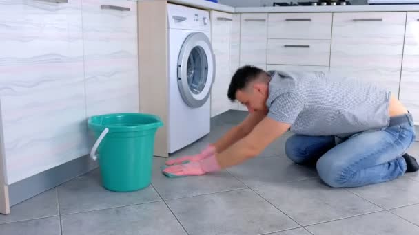 Homem cansado infeliz em luvas de borracha lava e esfrega duro o chão na cozinha, sentado no chão, vista lateral . — Vídeo de Stock