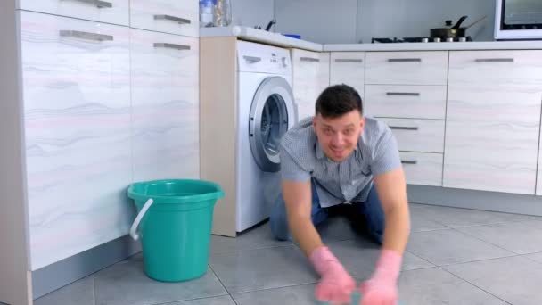 Hombre feliz con guantes de goma lava y frota duro el suelo en la cocina, sentado en el suelo y mirando a la cámara . — Vídeo de stock