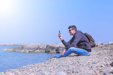 Onun akıllı telefon adam görüşmeler görüntülü sohbet tatilde deniz taş plajda oturur.