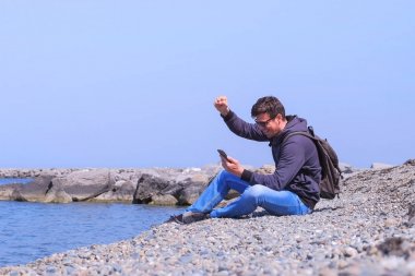Man gezgin deniz plajında oturur akıllı telefon büyük haber almak hakkında mutlu.