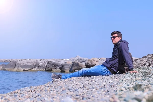 Adam turist gezgin taş plaja gelir oturur ve denize bakar.