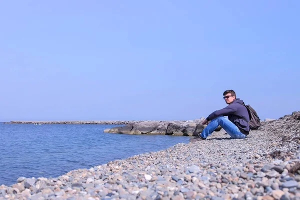 Молодой человек путешественник сидит на каменном пляже и смотрит на море на весеннем отдыхе . — стоковое фото