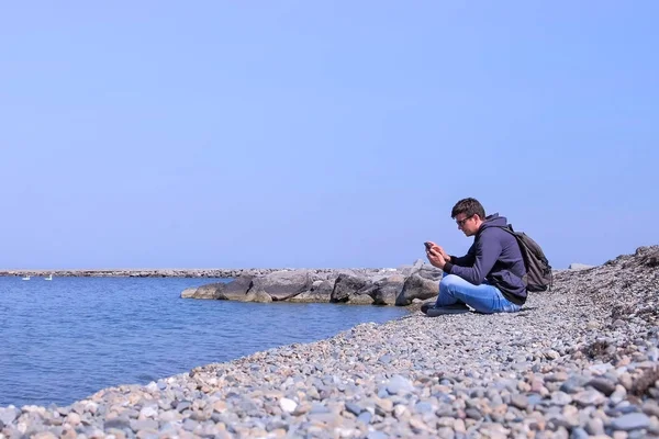 Mužíček sedí na mořské kamenné pláži a prochází smartphone, pohled z boku. — Stock fotografie