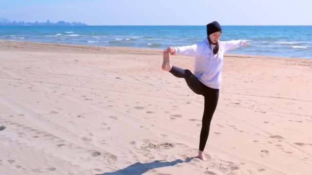 Γυναίκα κάνει γιόγκα στην παραλία της θάλασσας υψώνει το πόδι της και κρατά το χέρι σε ισορροπία πόζα. — Αρχείο Βίντεο