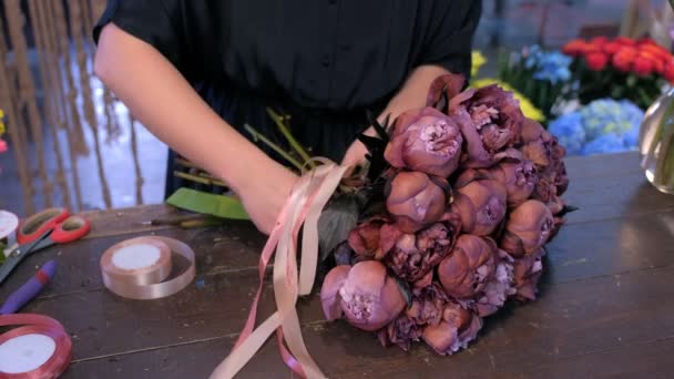 Woman florist ties stems by ribbons on bouquet of brown peonies in flower shop. — Stock Video