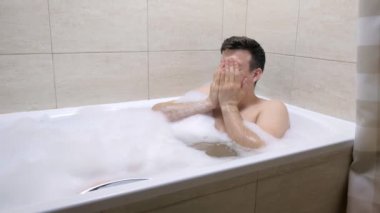 Banyo köpükte banyoda oturan yetişkin adam, elleri ile yüz mendiller.