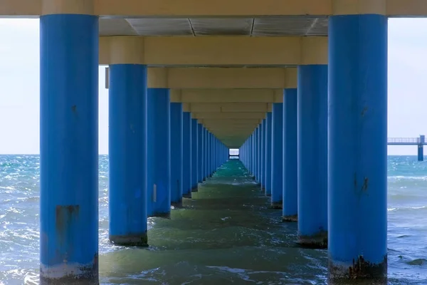 Sea under pier among parallel blue columns making narrow corridor in water.