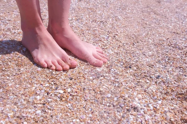 海辺の貝殻の砂浜で足をマッサージする裸足の女の子旅行者. — ストック写真