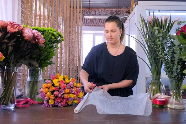 Mulher florista trabalha corta folha de papel para embalar buquê de flores na loja florística . — Fotografia de Stock