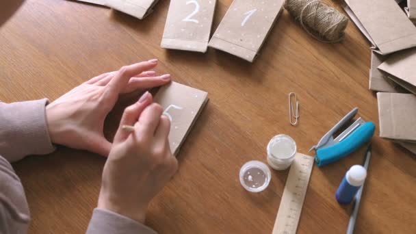 Woman makes christmas advent calendar for kids. Writes a number on the bag by brush and white paint. Close-up hands. — Stock Video