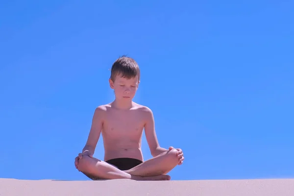 Niño niño está meditando en la pose de loto en la playa de arena en el fondo del cielo azul . — Foto de Stock